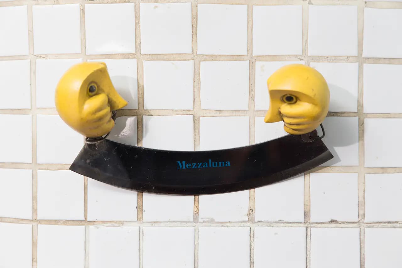 A curved mezzaluna chopping knife with two carved yellow handles mounted on a white tiled wall, the restaurant's name printed on the blade