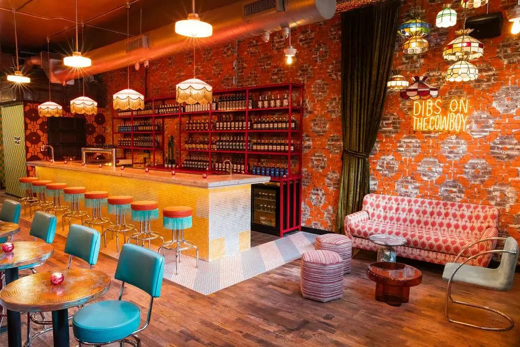 The front bar at Velvet Cowboy with orange-and-turquoise stools, fringed lamps and a neon sign reading Dibs on the Cowboy