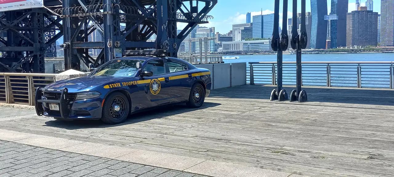 A dark blue New York State Police Dodge Charger with gold stripes parked on a waterfront pier with the Manhattan skyline behind it