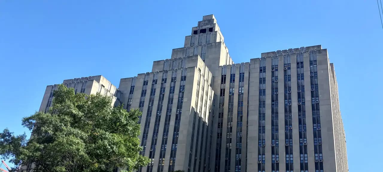 The tall Art Deco limestone tower of the Manhattan Criminal Court at 100 Centre Street against a clear blue sky, with a tree in the foreground