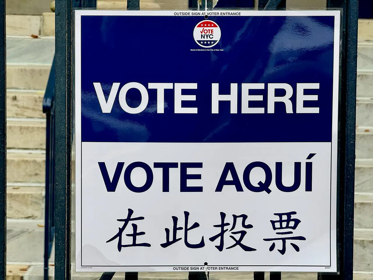 A blue and white 'Vote Here / Vote Aquí' sign in English, Spanish and Chinese from the NYC Board of Elections on a fence outside a poll site