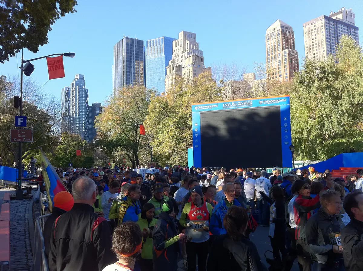 Runners wrapped in jackets gather near the New York City Marathon finish area in Central Park under autumn trees and a big video screen, with Central Park South towers behind