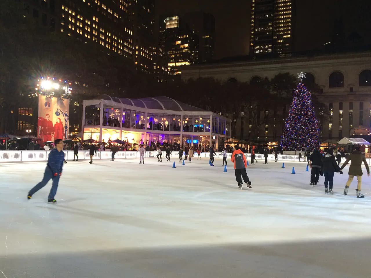 Skaters on the rink at Bank of America Winter Village in Bryant Park at night, with a lit Christmas tree and the back of the New York Public Library's main building behind them